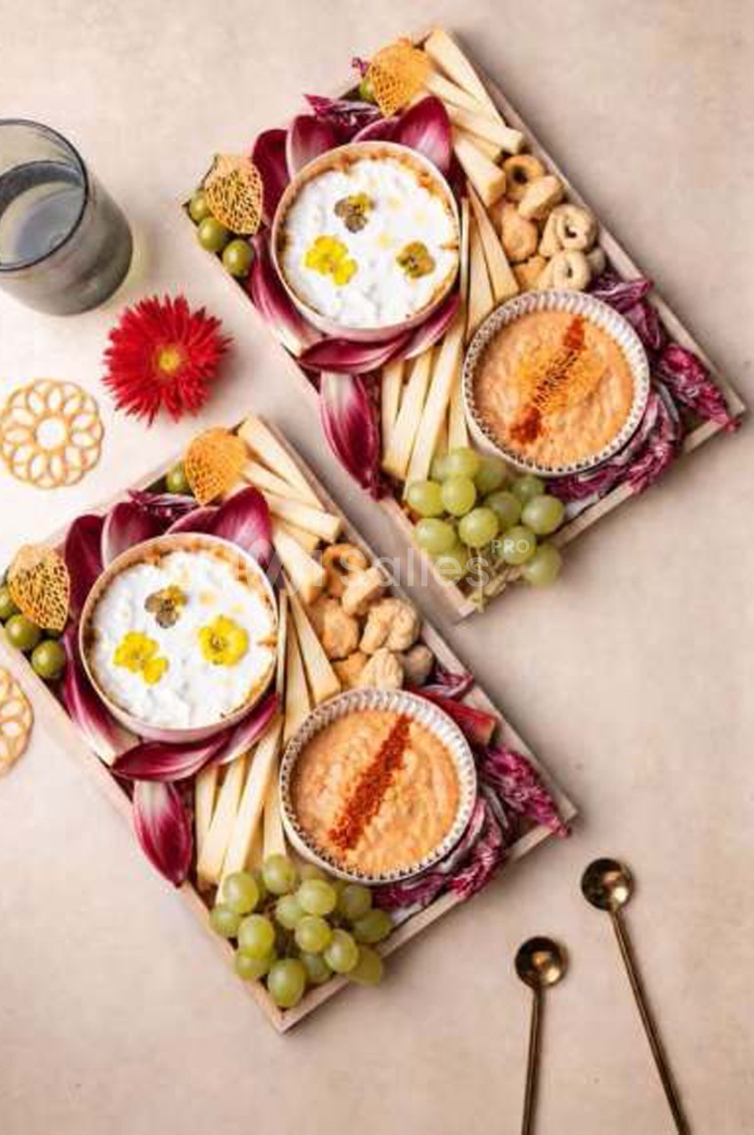 Deux planches apéritives en bois disposées en diagonale, avec dips blancs et orangés garnis de paprika, crudités, raisins verts, fromage en bâtonnets et fleurs comestibles.
