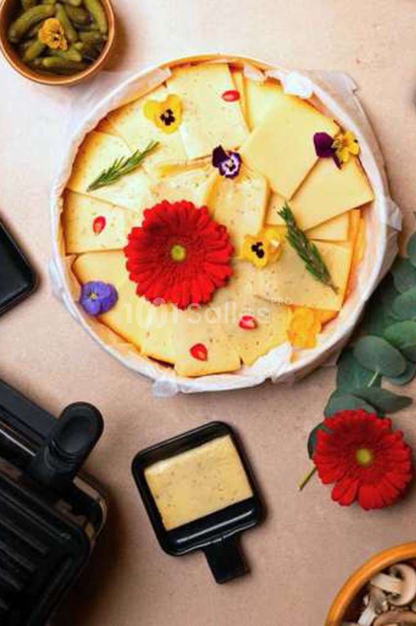 Plateau rond de fromage à raclette en tranches, décoré de fleurs comestibles rouges et jaunes et de brins de romarin, à côté d'une coupelle de fromage fondu.