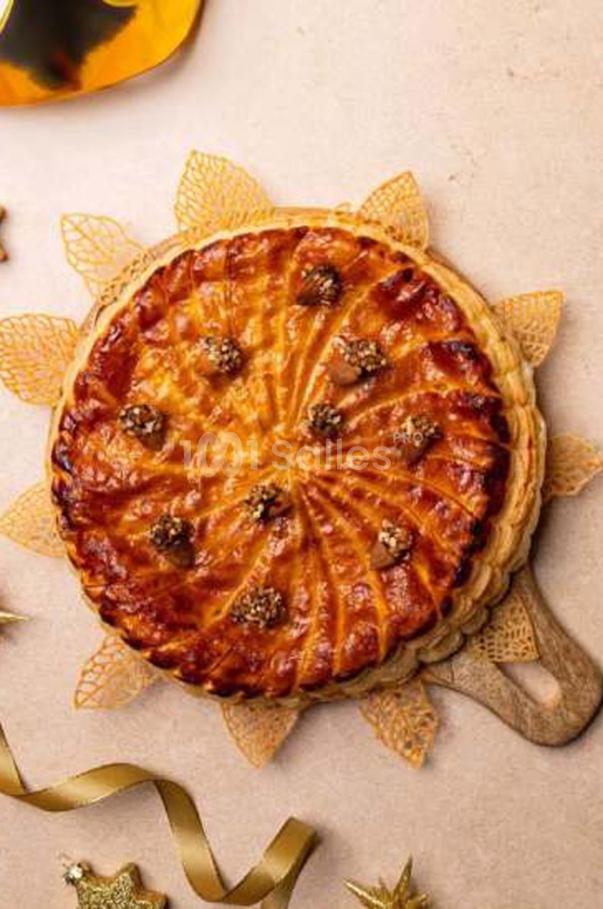 Galette des rois dorée à la frangipane, décorée de rosaces en pâte et de noix ou amandes disposées en cercle, posée sur une planche en bois avec des ornements dorés. Wait, I must check: "noix ou amandes" - the decorations look like nuts