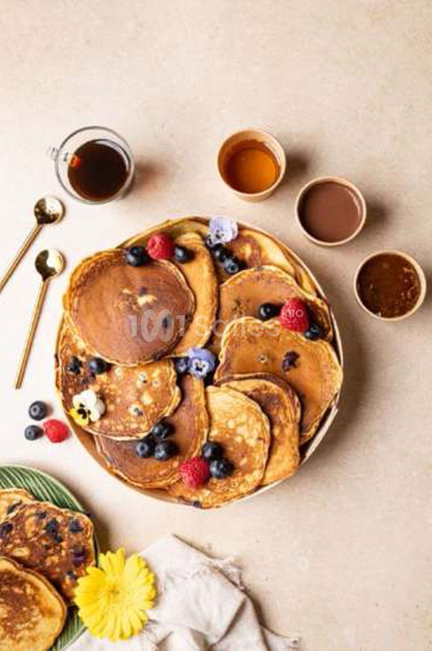 Pancakes dressés en pile sur un plateau rond, garnis de myrtilles et de framboises, accompagnés de petits pots de sirop et de sauces.