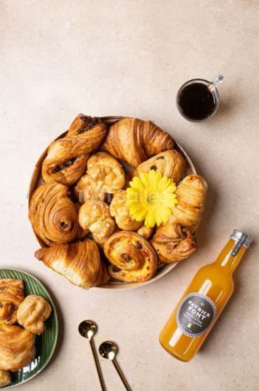 Plateau de viennoiseries (croissants, pains au chocolat, chouquettes) disposé sur une assiette beige, accompagné d'un jus d'orange en bouteille et de cuillères dorées sur une table claire.
