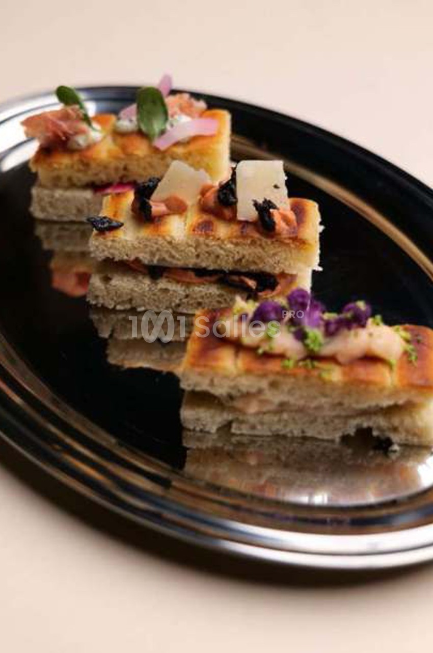 Trois carrés de focaccia dorée empilés sur un plateau ovale noir, garnis de jambon cru, de copeaux de fromage, d'olives noires et de fleurs violettes.