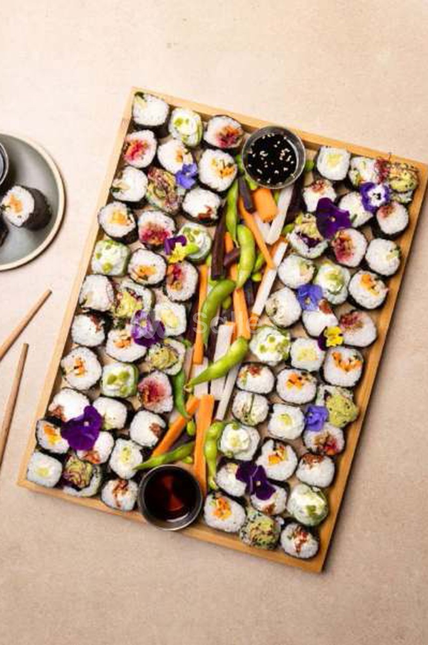 Plateau rectangulaire en bois garni de nombreux makis ronds à la fleur violette et aux légumes colorés, avec bols de sauce soja et bâtonnets de carottes et de haricots edamame.