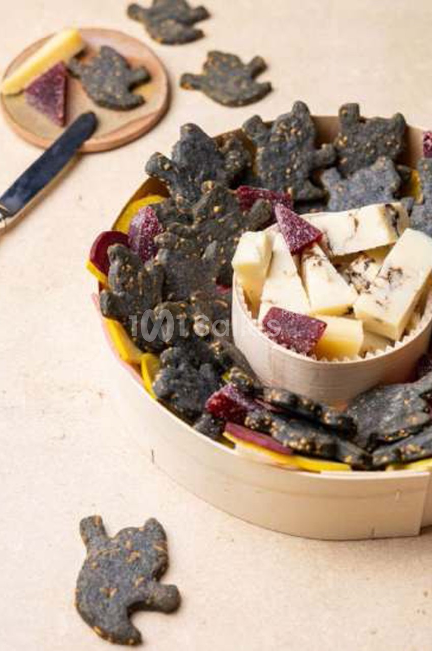 Biscuits sablés noirs en forme de chats, parsemés de graines, disposés avec des cubes de fromage blanc et des morceaux de betterave dans une coupelle en bois.