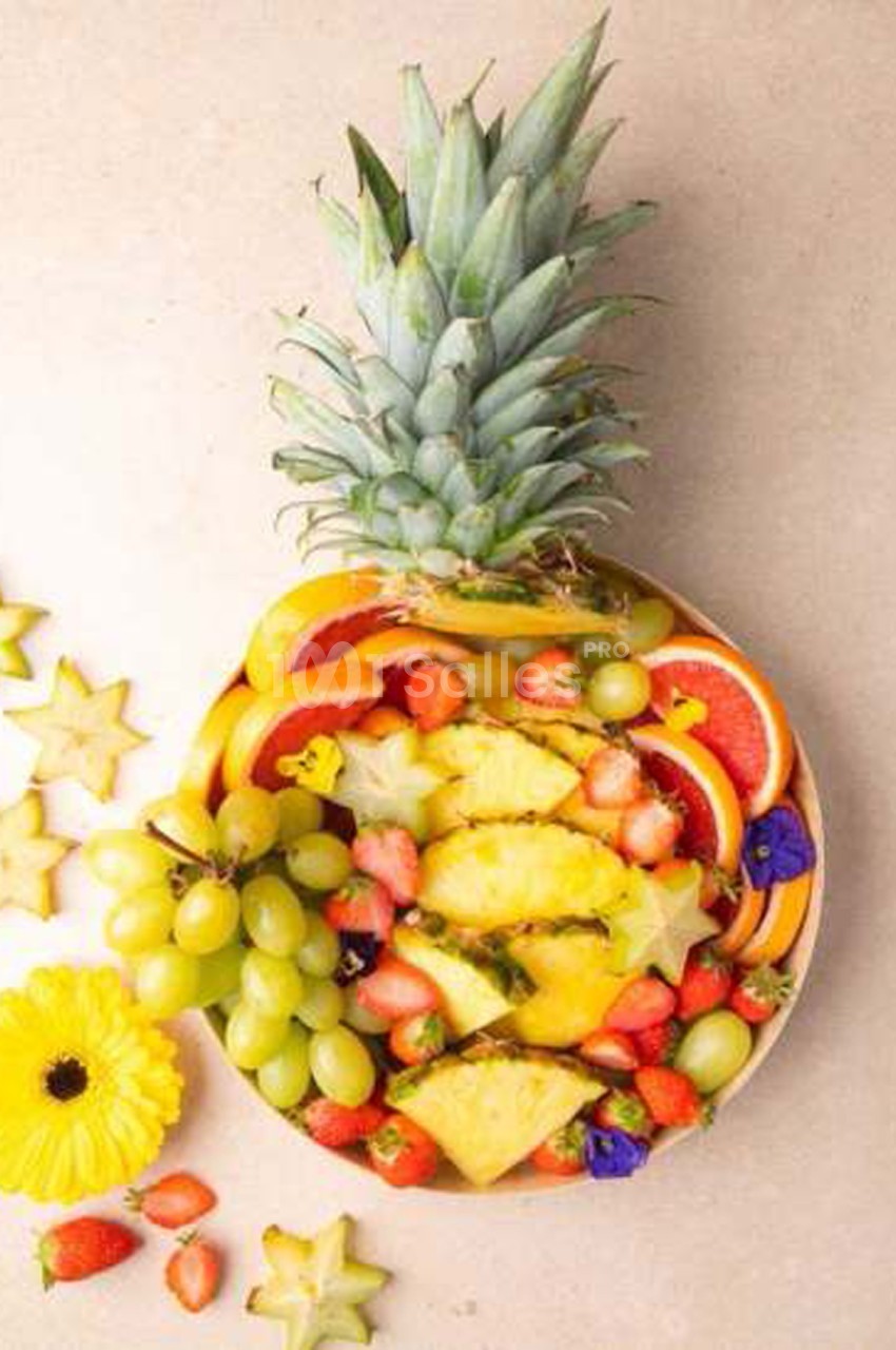 Ananas évidé rempli de tranches de fruits : pamplemousse, orange, fraises, raisins verts, caramboles et ananas, disposés sur fond clair.