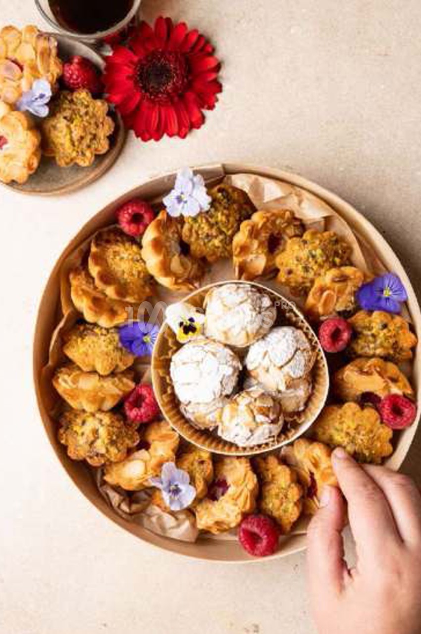 Plateau rond de petits gâteaux dorés aux amandes et aux pistaches, décorés de framboises et de fleurs violettes, avec une coupelle centrale de biscuits poudrés au sucre glace.