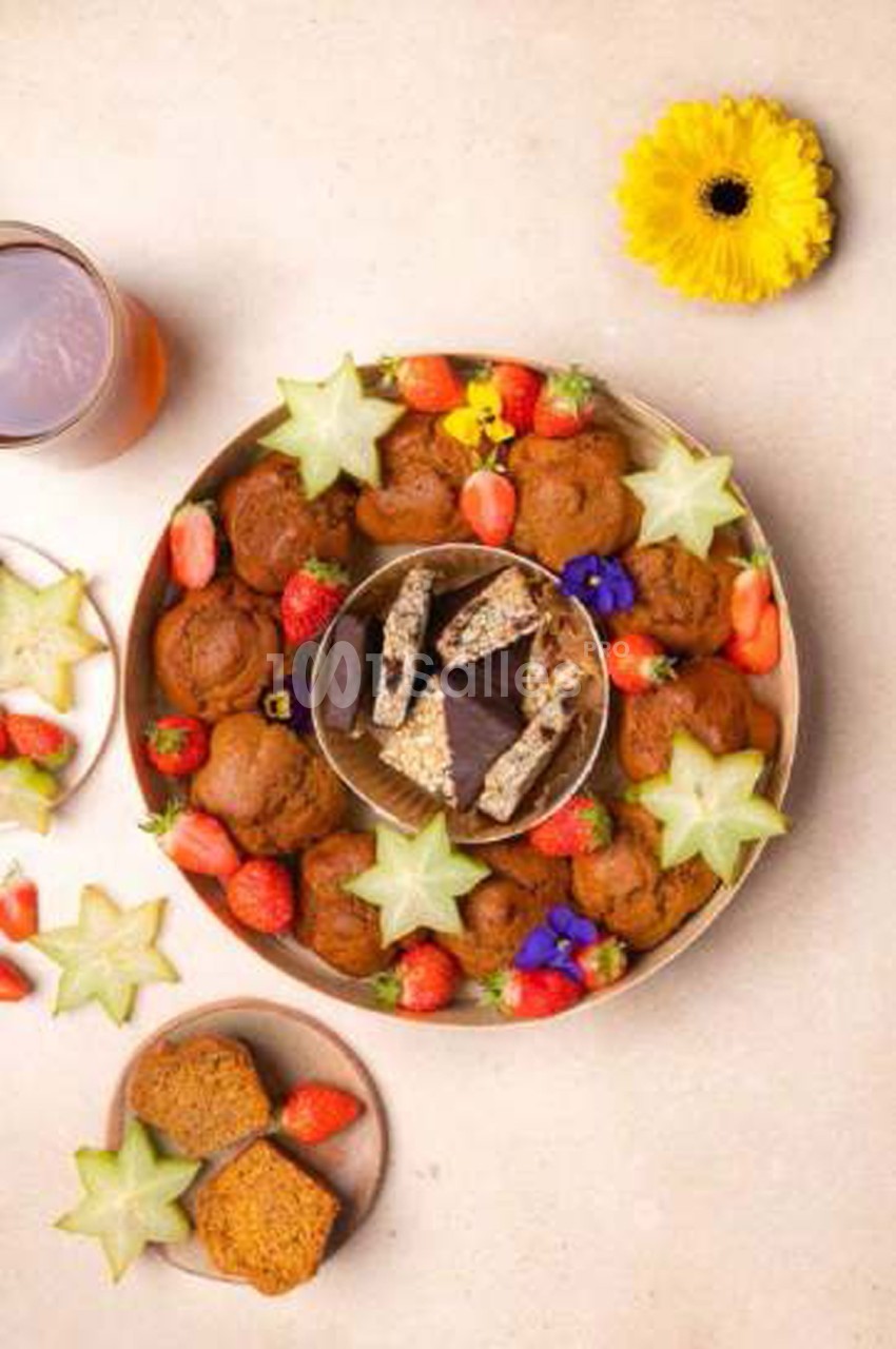Plateau rond de petits gâteaux bruns, fraises, caramboles en étoile et fleurs comestibles, avec des barres aux éclats de noix dans une coupelle au centre.