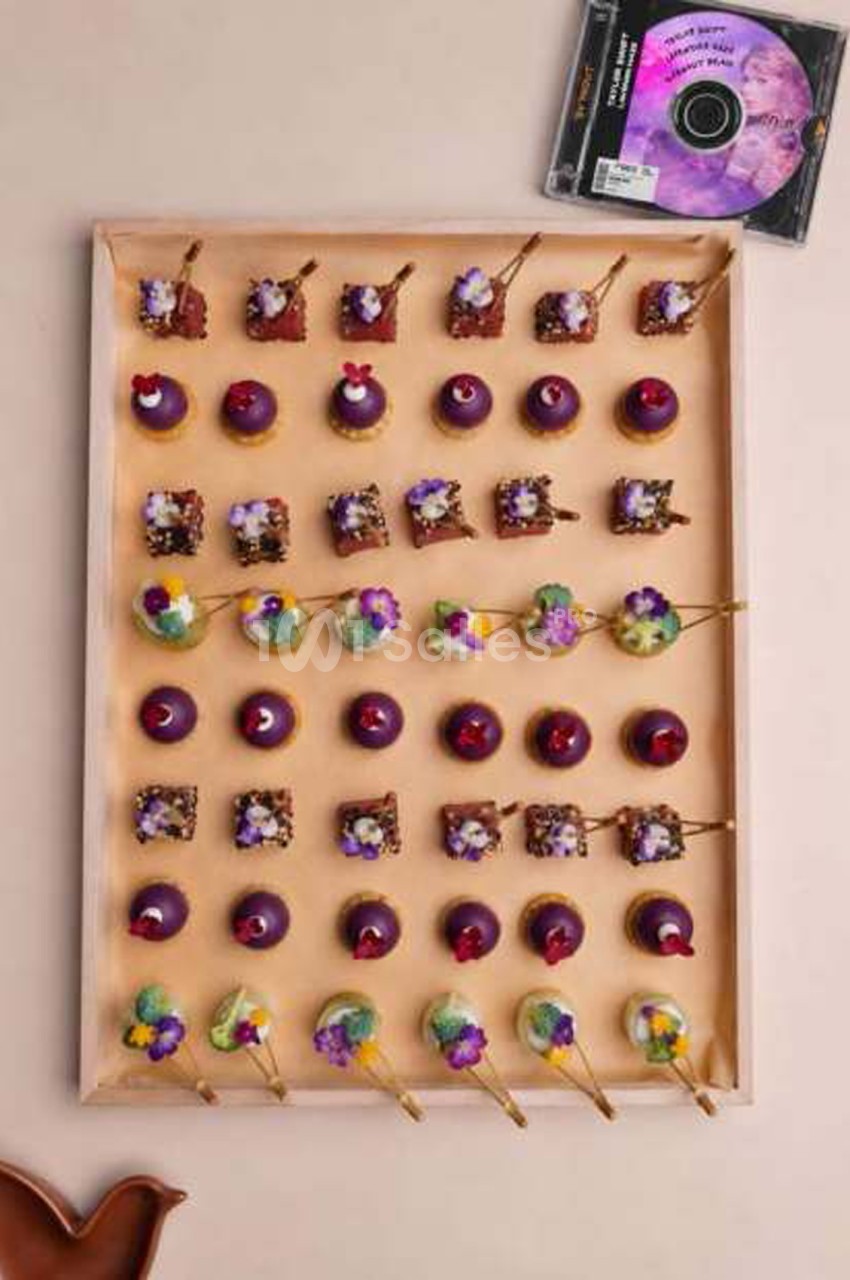 Plateau en bois de petites pâtisseries violettes disposées en grille, décorées de fleurs comestibles et de pics dorés.