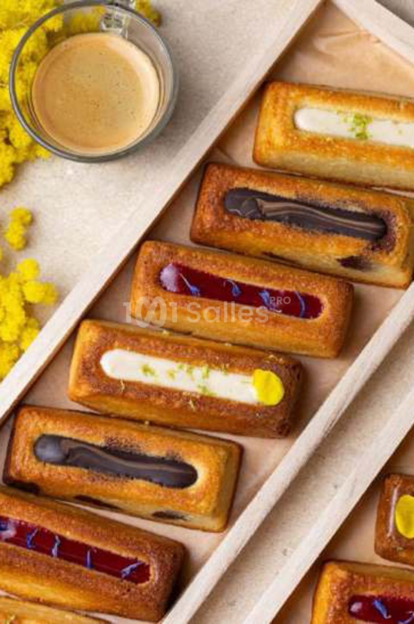 Financiers allongés disposés en diagonale dans un plateau en bois, garnis de crème blanche, de ganache chocolat, de confiture rouge et de pâte de fruit jaune, avec une tasse de café en haut à gauche. Let me check length: roughly 165 characters, too long.