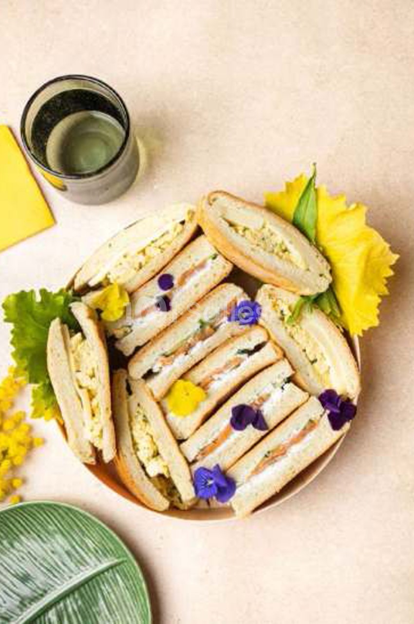 Plateau de sandwiches au pain blanc garnis d'œufs et de saumon fumé, disposés en éventail et décorés de fleurs violettes.