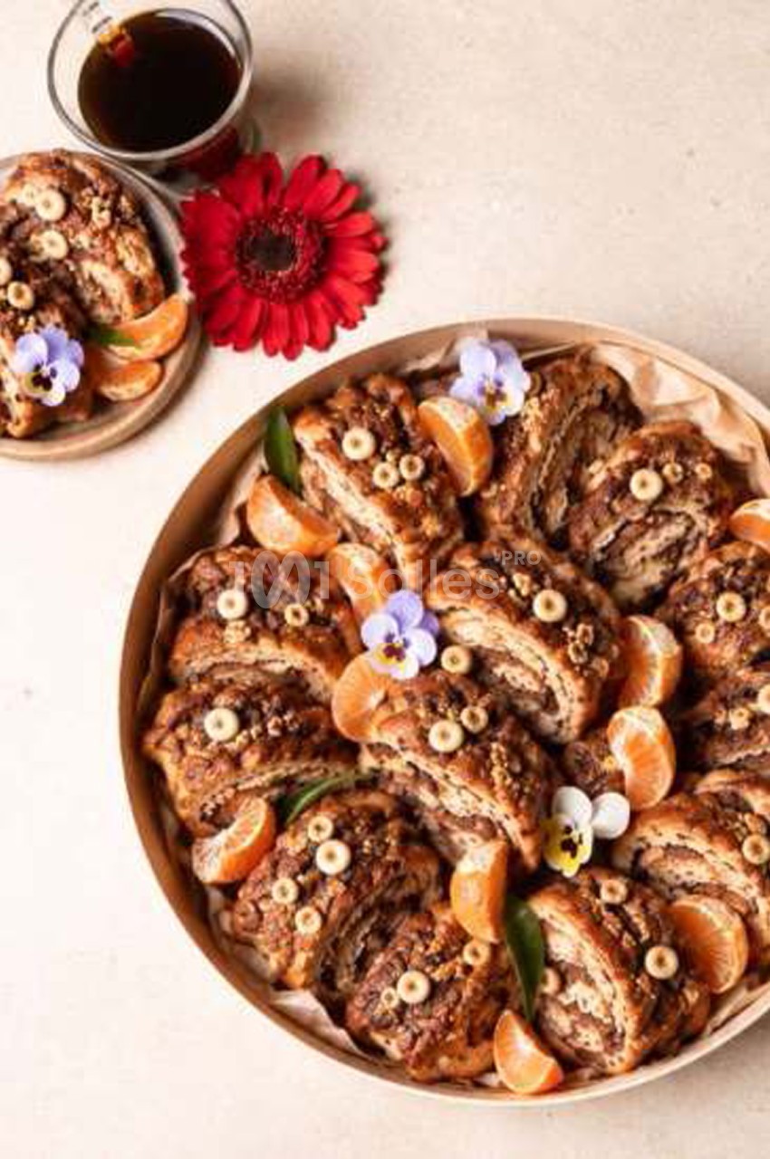 Plateau rond de viennoiseries marbrées garnies de pépites et de noix, décoré de quartiers de mandarine, de fleurs violettes et de feuilles vertes, avec une tasse de café.
