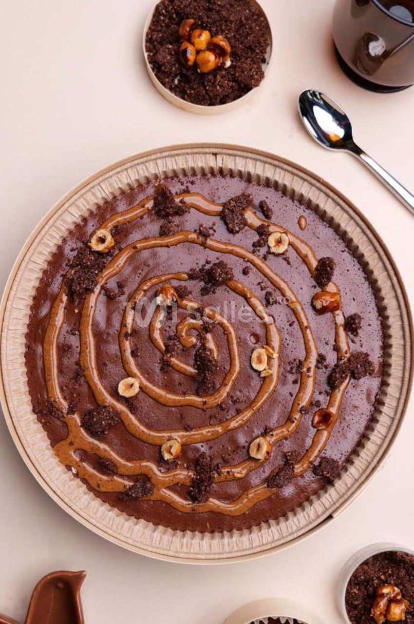 Tarte ronde au chocolat garnie d'un décor en spirale de caramel à la noisette, de noisettes caramélisées et de crumble au cacao, vue de dessus sur fond clair.