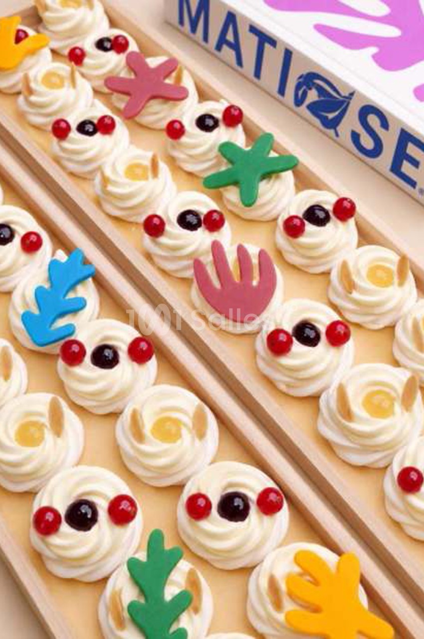 Petites pâtisseries en rosaces de crème blanche décorées de groupes de cerises, de raisins noirs et de formes colorées en pâte d'amande, disposées en rangées sur des plateaux en bois.