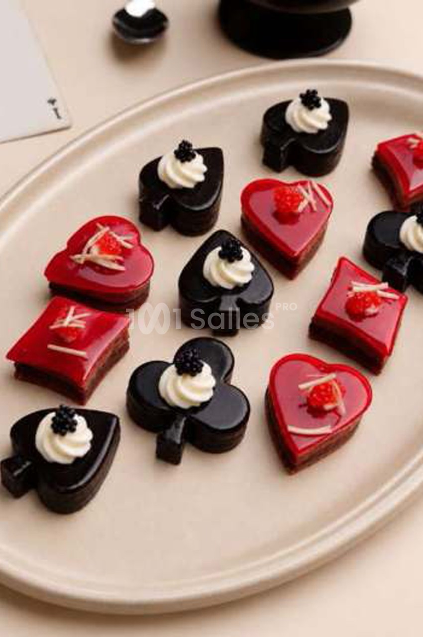 Petits gâteaux en forme de cœurs, piques, trèfles et carreaux, glacés en rouge ou en noir, garnis de crème et de perles noires. Let me check length: roughly 125 characters? Count: "Petits gâteaux en forme de cœurs, piques, trèfles et carreaux, glacés en