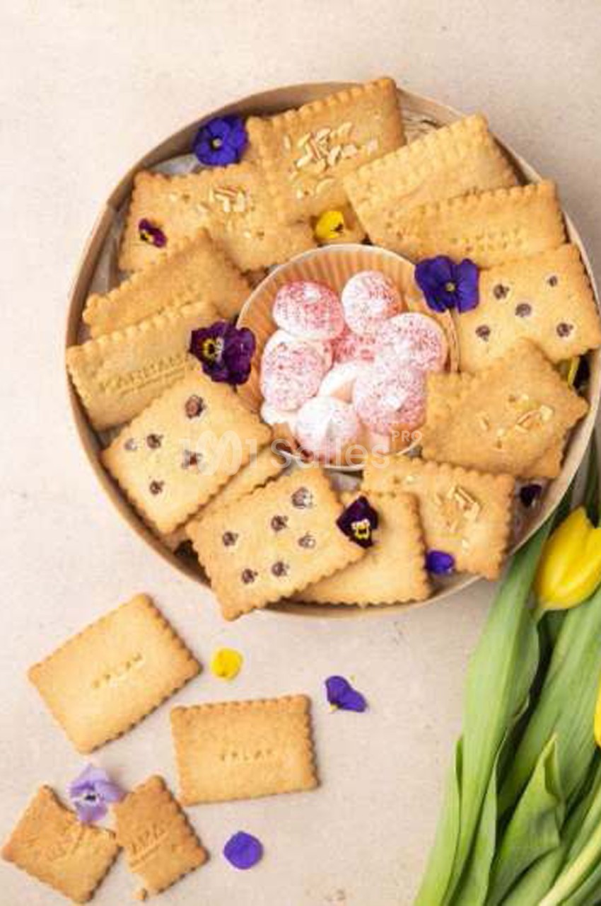 Plateau rond de biscuits sablés, dont certains avec des pépites de chocolat ou des amandes effilées, décoré de fleurs violettes comestibles et entourant une coupelle de meringues roses. Count characters: roughly 180 characters, too long. Need ≤125.