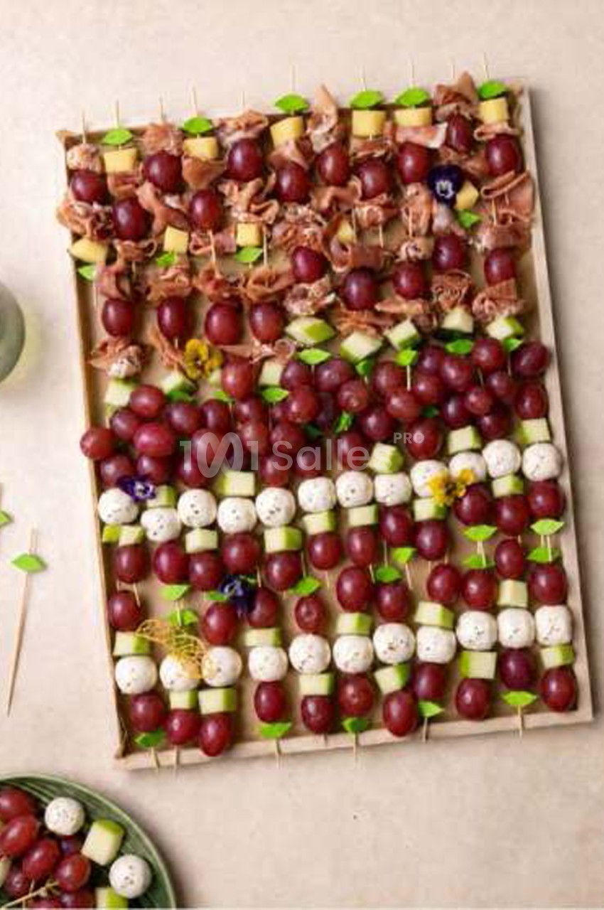 Plateau de piques apéritives disposées en rangées : raisin rouge, dés de melon, boules de mozzarella, fines tranches de jambon cru et feuilles vertes. Let me check constraints: one sentence, under 125 chars? Count: "Plateau de piques apéritives disposées