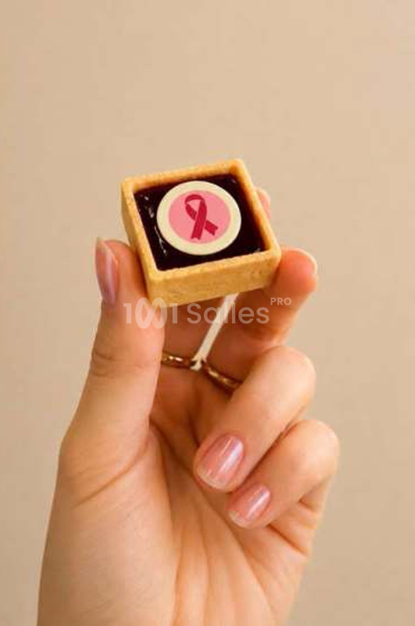 Main tenant une petite tartelette carrée garnie d'une préparation sombre, ornée d'un disque rose à ruban rouge.