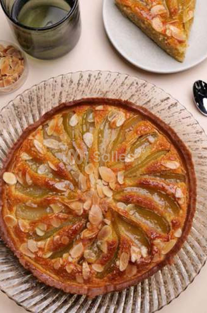 Tarte feuilletée aux poires et amandes effilées, disposée en rosace sur une assiette en verre, à côté d'une part servie. Count: roughly 120 characters. Fine.