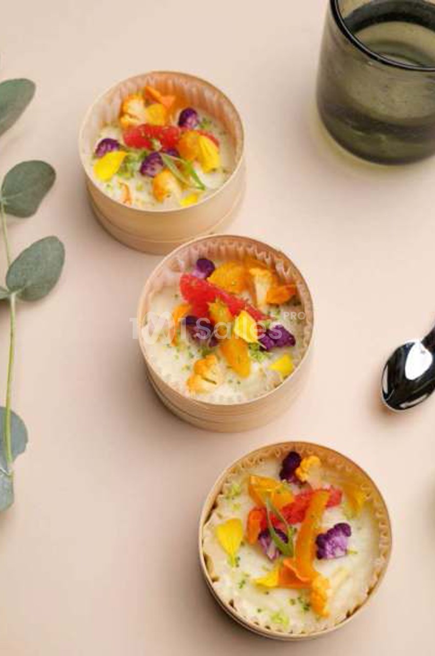 Trois petites coupelles en bois contenant une crème blanche garnie de légumes colorés (carotte, chou-fleur violet, pétales jaunes) et de fleurs comestibles, posées sur une surface beige.