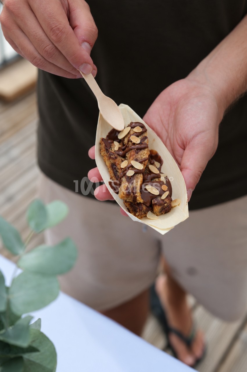 Personne tenant dans sa main une coupelle en bois contenant une part de gâteau nappée de chocolat et parsemée d'amandes effilées, avec une petite cuillère en bois. Let me check length: roughly 165 characters, too long. Need ≤125. Shorter: "Main tenant
