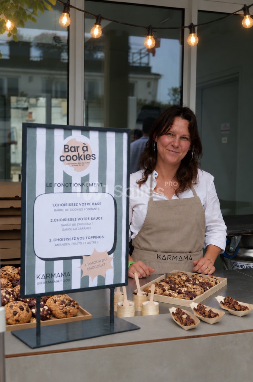 Femme souriante derrière un comptoir de bar à cookies, avec une pancarte d'instructions et des cookies et desserts sur plateau en bois, sous des guirlandes lumineuses. Count characters: roughly 170? Let's count more carefully. Need ≤125. Shorten. "Femme