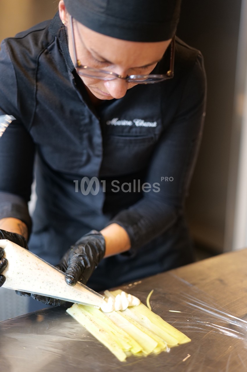 Personne en tenue de cuisine noire et gants garnissant de crème des bandes de poireau sur un plan de travail inox.