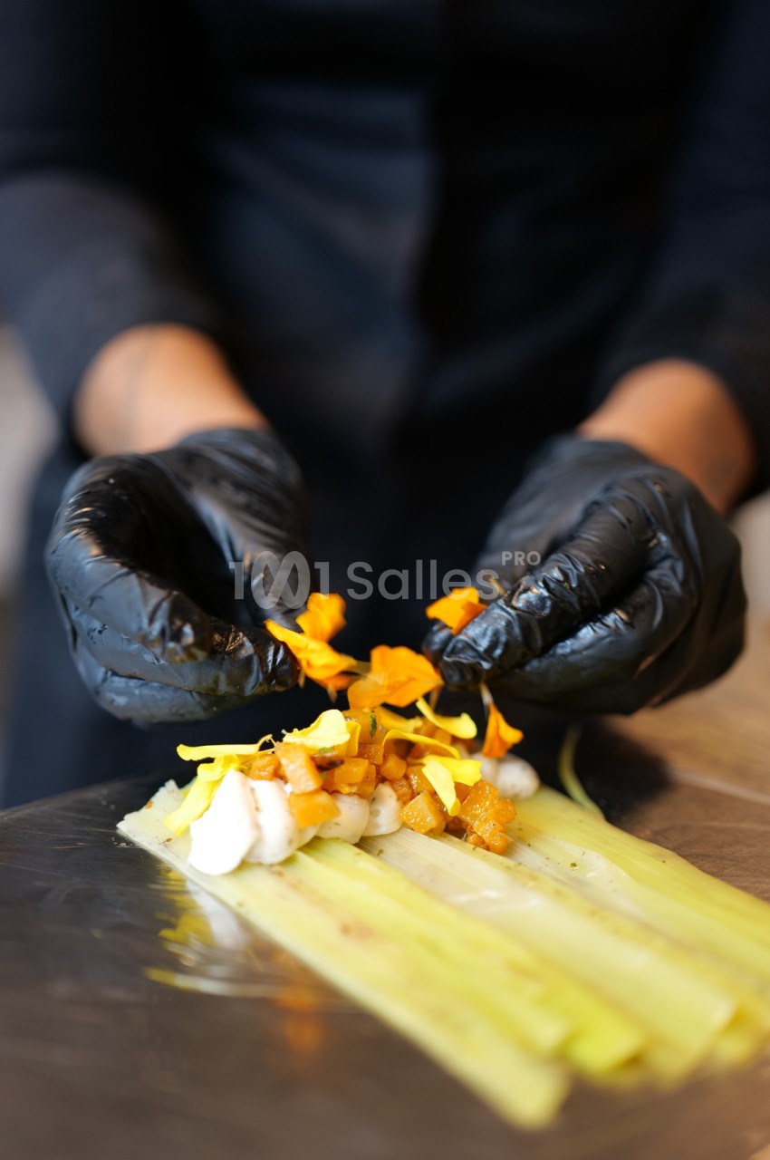 Mains gantées de noir dressant sur une feuille de poireau une préparation de dés orangés, de crème blanche et de pétales de fleurs jaune-orangé.