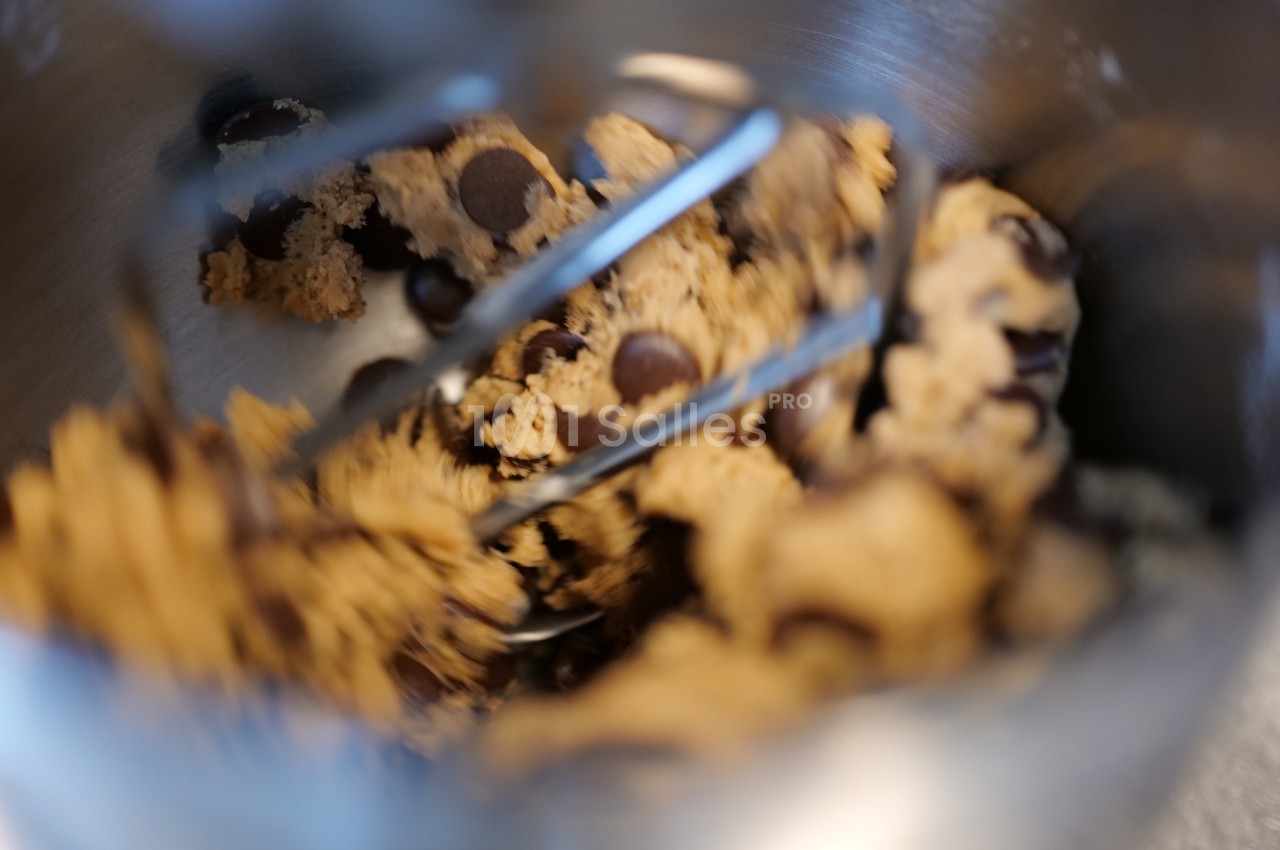Pâte à cookies brune aux pépites de chocolat, malaxée dans un bol en inox par les batteurs d'un robot de cuisine.