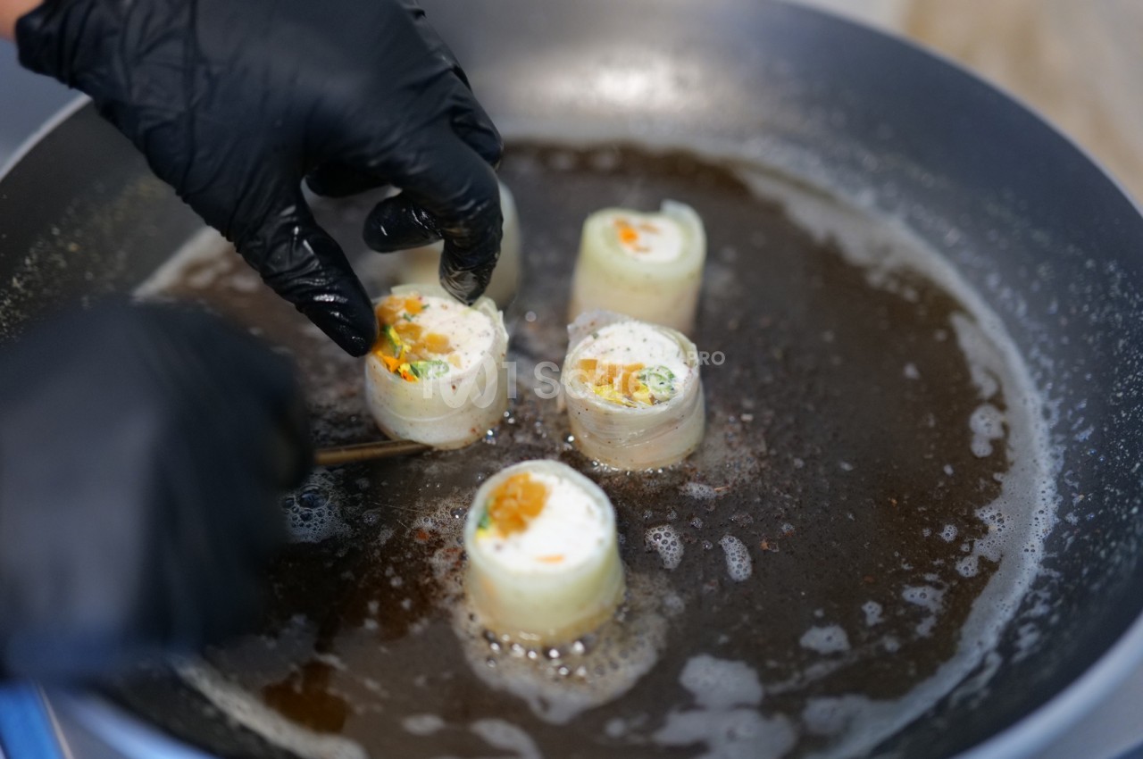Mains gantées de noir disposant des rouleaux farcis dans une poêle chaude, garnis de crème et de dés orangés.