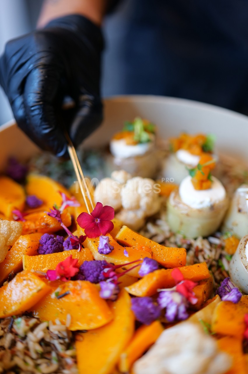 Une main gantée de noir pique une fleur comestible violette sur un plat de légumes rôtis : tranches de potiron, chou-fleur violet et blanc, riz sauvage et bouchées garnies de fromage frais.