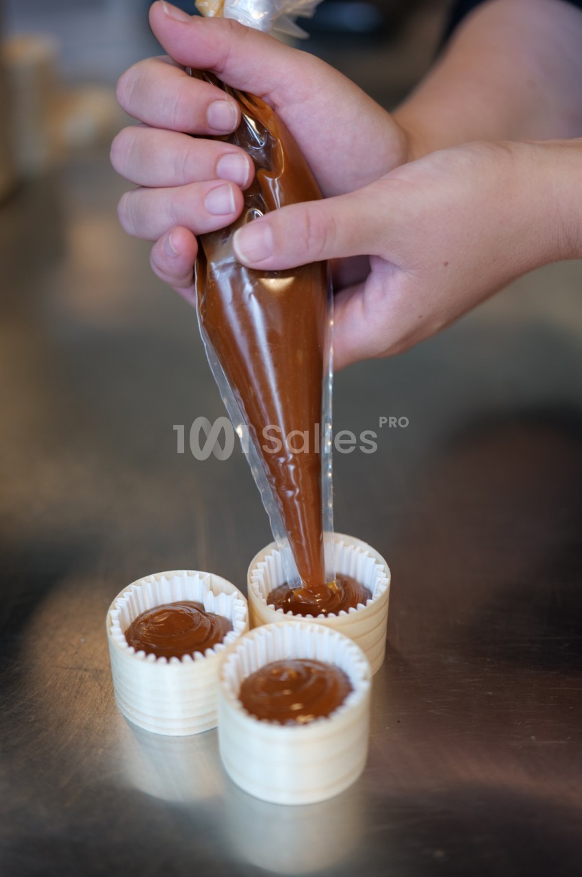 Mains tenant une poche plastique remplie de caramel qui garnit de petites caissettes blanches posées sur un plan de travail en inox.