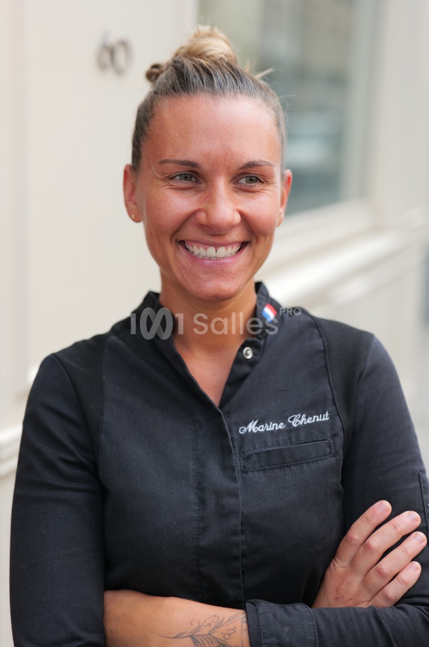 Femme souriante en veste de cuisine noire, bras croisés, cheveux relevés en chignon, devant un mur clair en extérieur.
