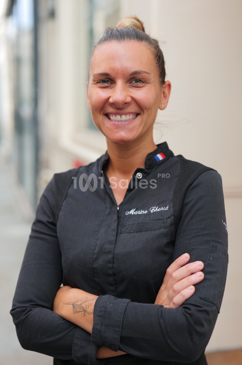Femme souriante aux cheveux attachés en chignon, bras croisés, portant une chemise noire brodée d'un prénom et d'un petit drapeau français au col.