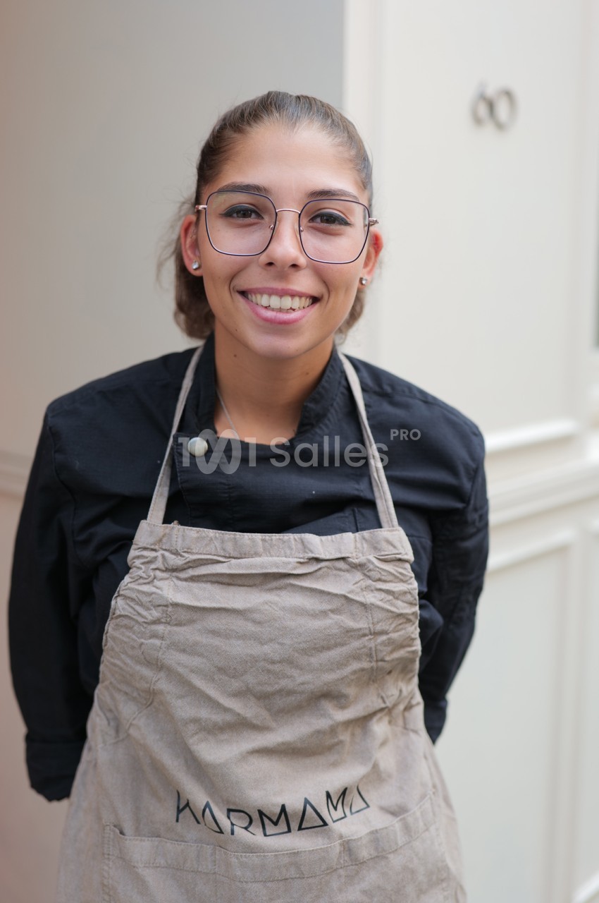 Jeune femme souriante aux lunettes fines, en veste noire et tablier beige, posant devant un mur clair.