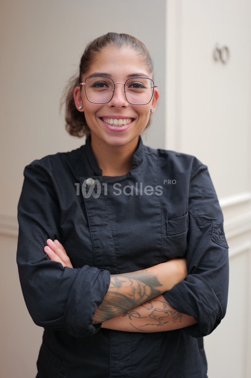 Femme souriante en veste noire de type cuisinier, bras croisés, devant un mur beige.