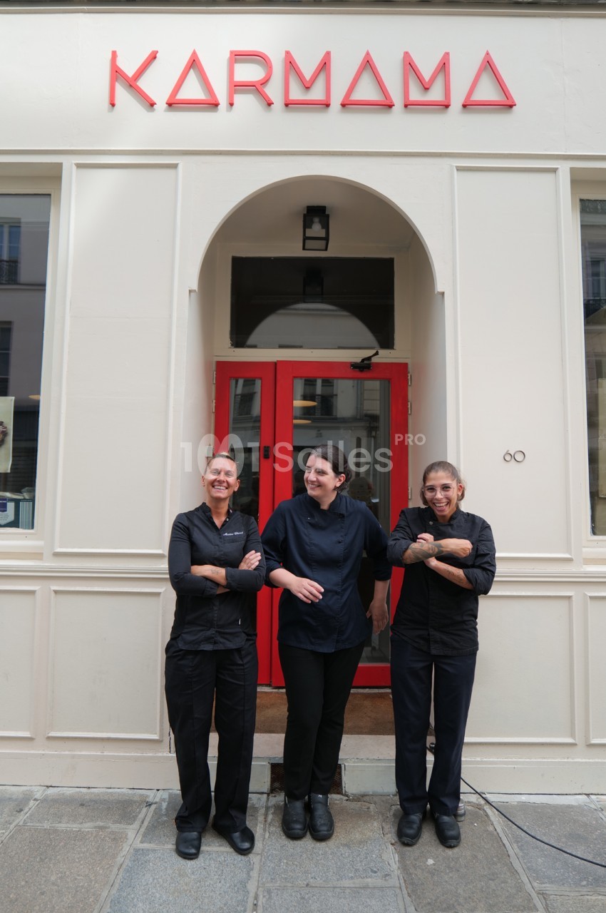 Trois femmes en tenue de cuisinier noire posent devant la façade blanche d'un établissement aux portes rouges, sous l'enseigne rouge KARMAMA. Wait, I must not transcribe text from the image (enseigne). So avoid "KARMAMA". Also the watermark "1001 Salles"