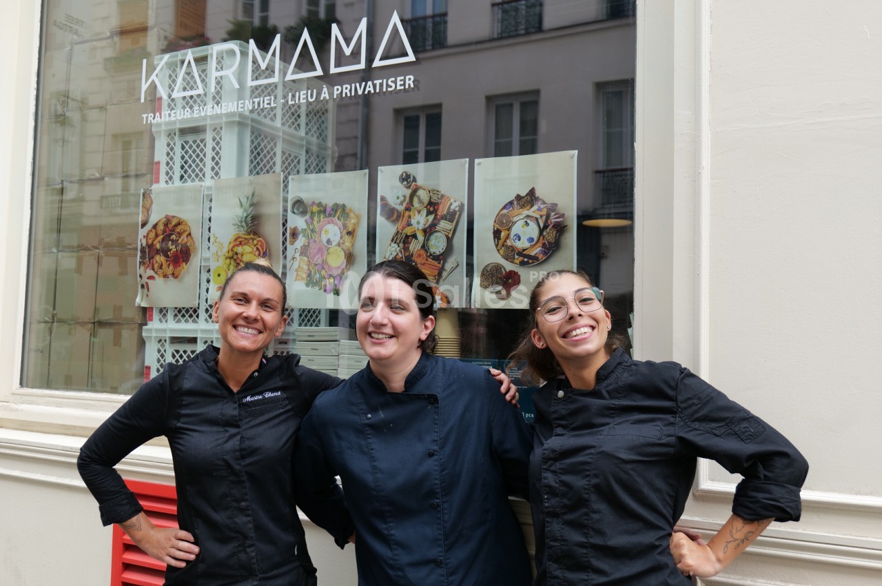 Trois femmes souriantes en tenue de cuisine noire posent devant une vitrine ornée de photos de plats colorés, dans une rue parisienne.