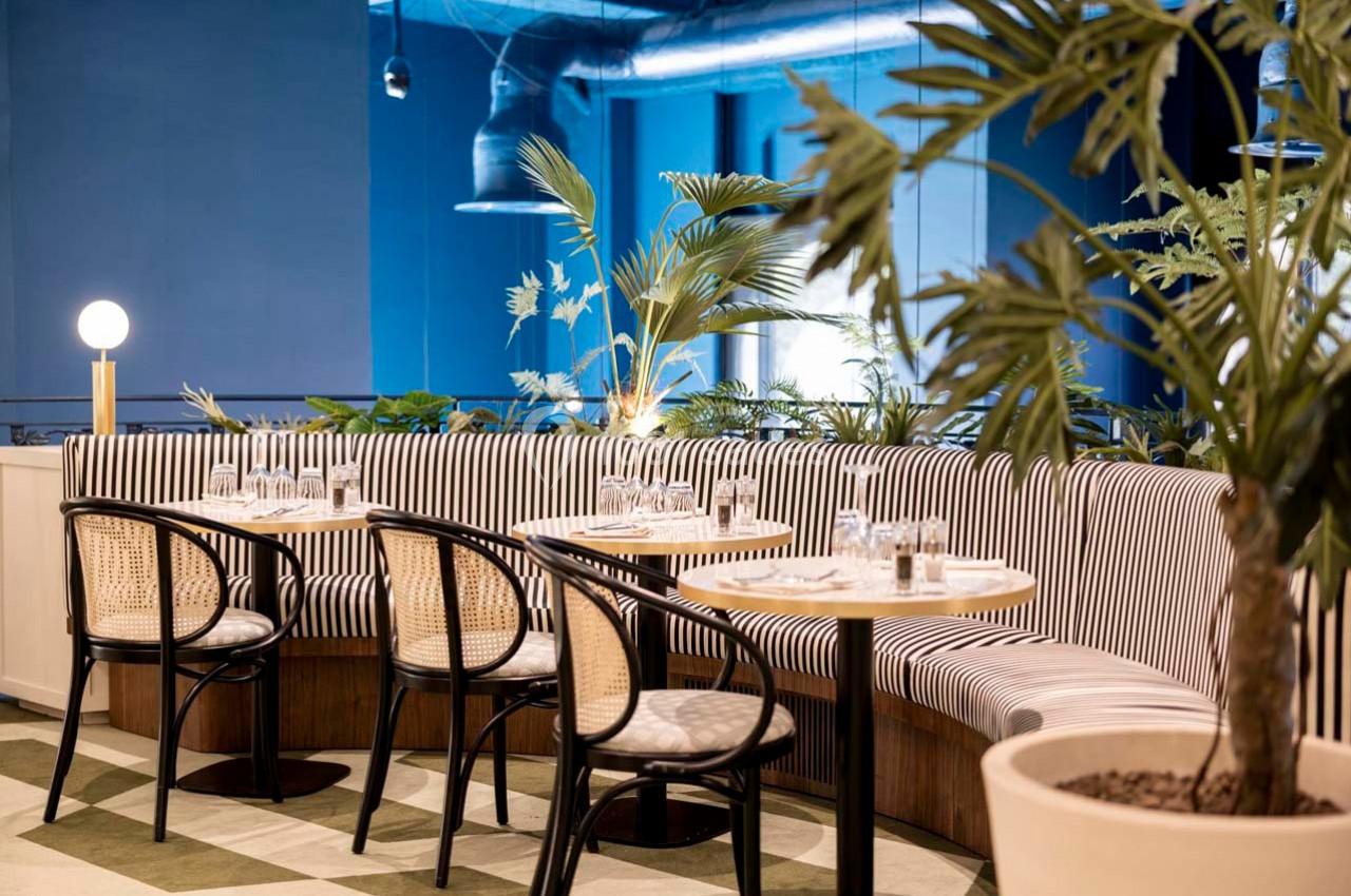 Banquettes rayées et chaises en rotin entourant des tables dressées dans un restaurant à la décoration végétale et moderne.