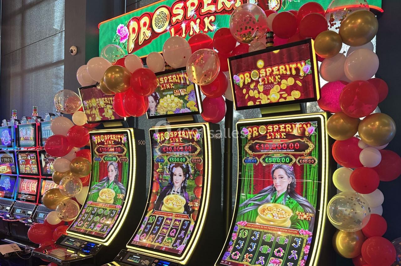 Machines à sous lumineuses décorées de ballons, affichant le jeu ’Prosperity Link’ dans un casino.