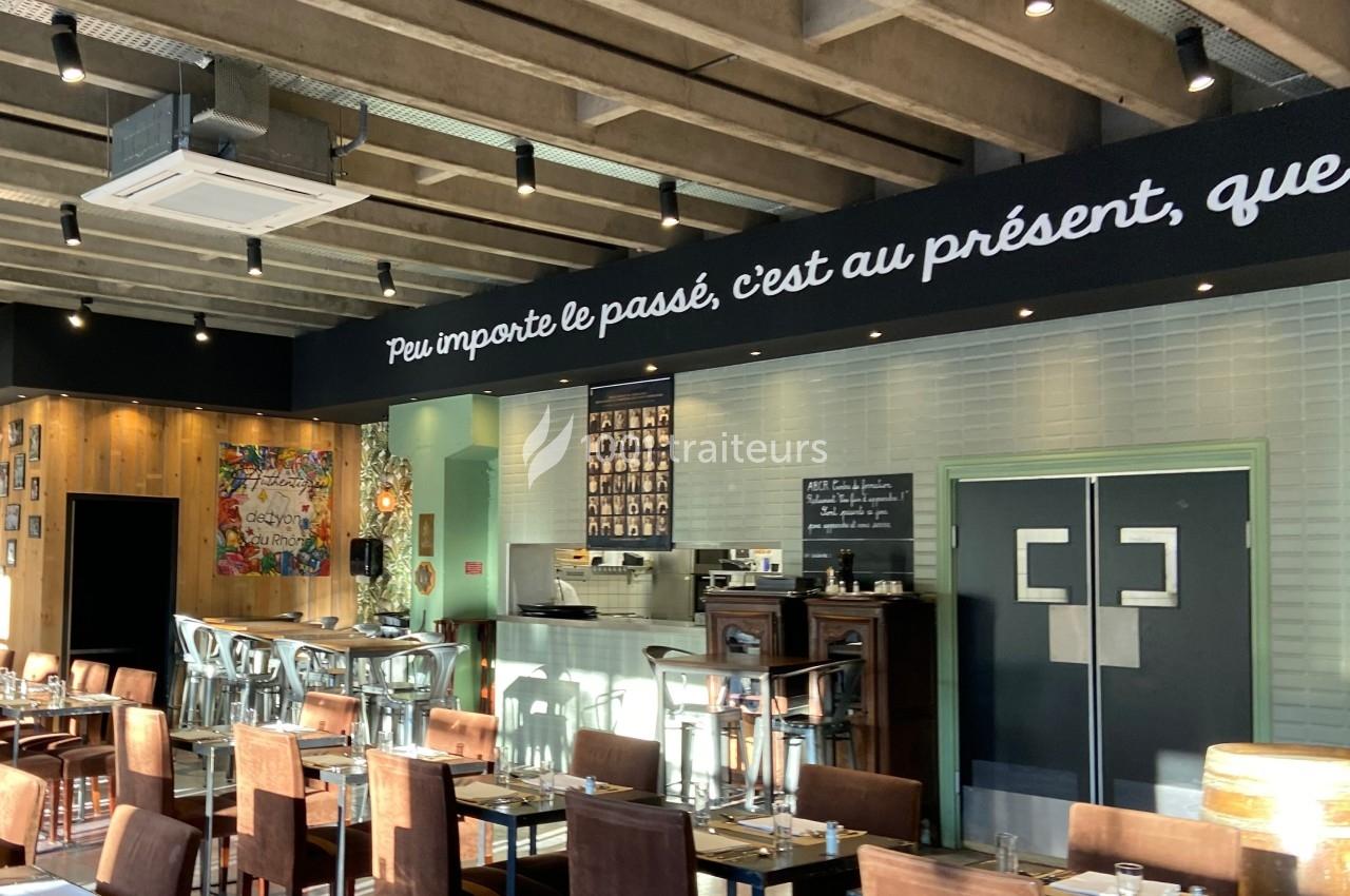 Salle de restaurant lumineuse avec tables dressées, mur décoré d'une citation et plafond en béton apparent.
