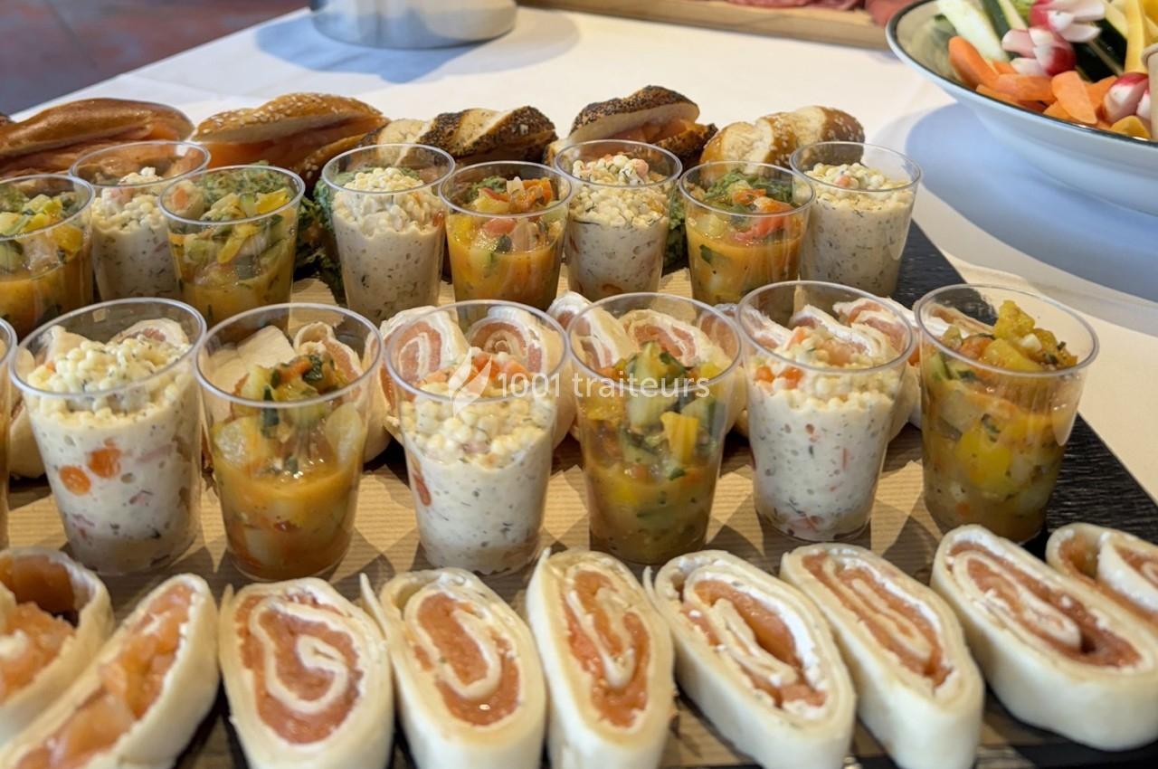 Plateau de verrines colorées et roulés au saumon disposés sur une table, accompagnés de pains et crudités.