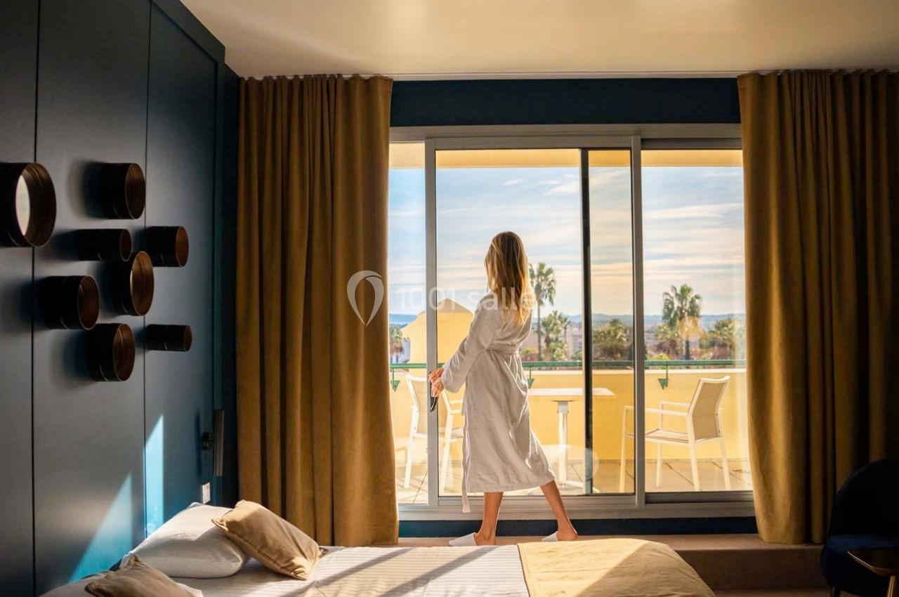 Location salle Hyères (Var) - Pasino de Hyères #12 Femme en peignoir regardant à travers une baie vitrée donnant sur une terrasse ensoleillée avec vue sur des palmiers.