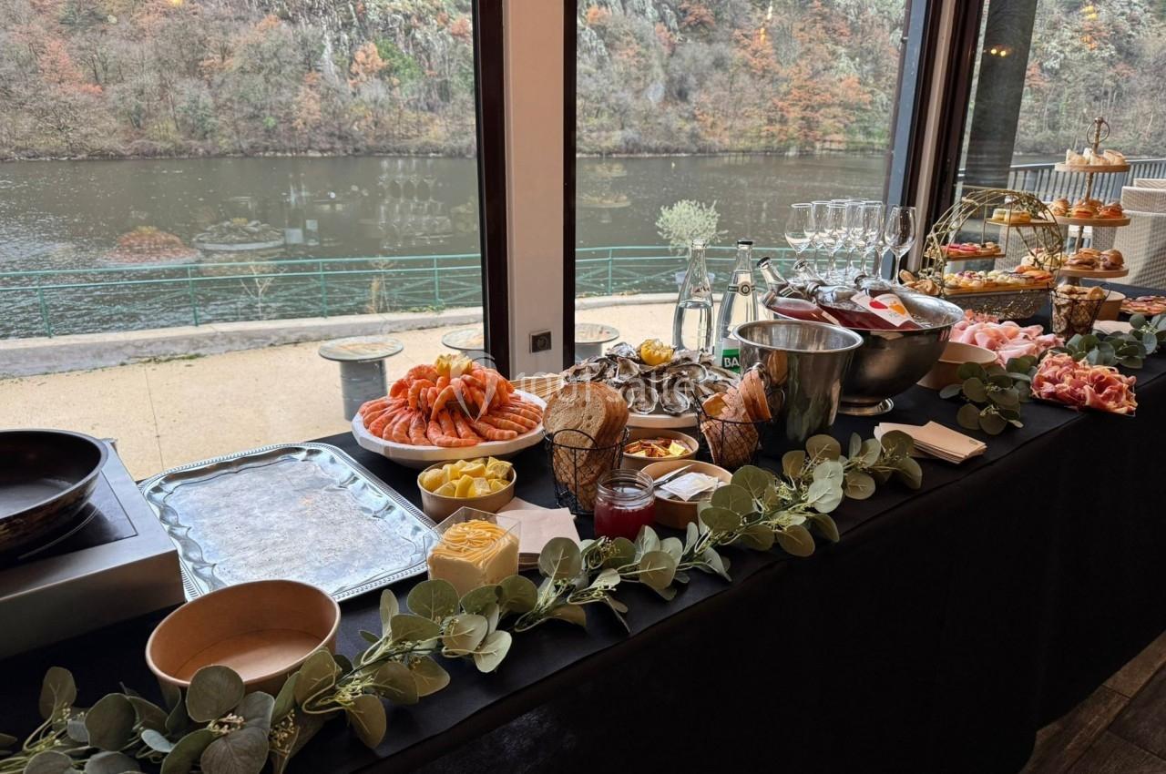 Buffet élégant avec viennoiseries, fruits et boissons, disposé près d'une baie vitrée donnant sur une rivière et des arbres.