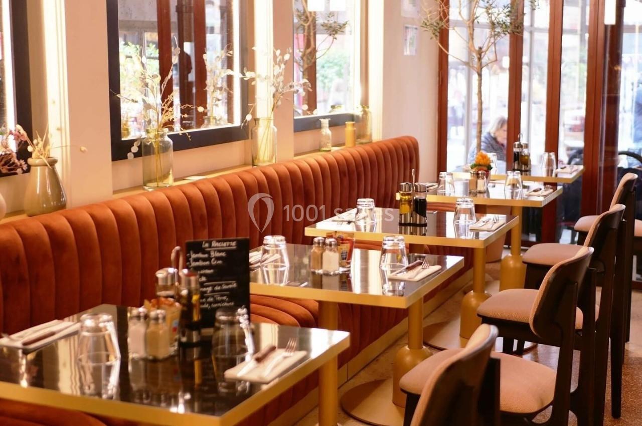 Salle de restaurant avec banquettes en velours orange, tables dressées et grandes baies vitrées laissant entrer la lumière.