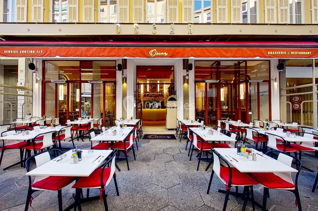 Terrasse d'un restaurant avec tables et chaises rouges et blanches, devant une façade vitrée et une enseigne rouge.