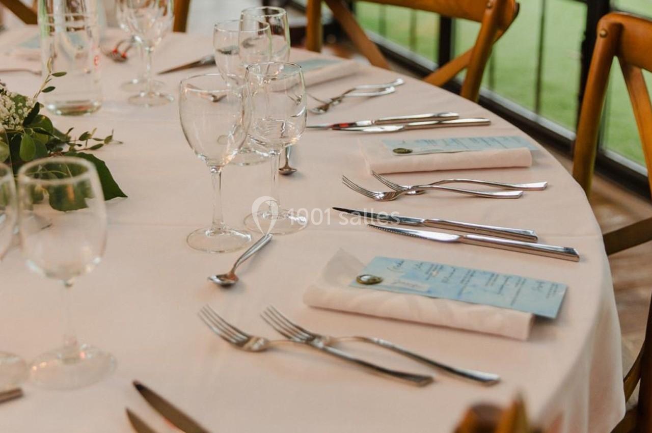 Table dressée avec nappes blanches, couverts, verres à vin, menus et décoration florale près d'une baie vitrée.
