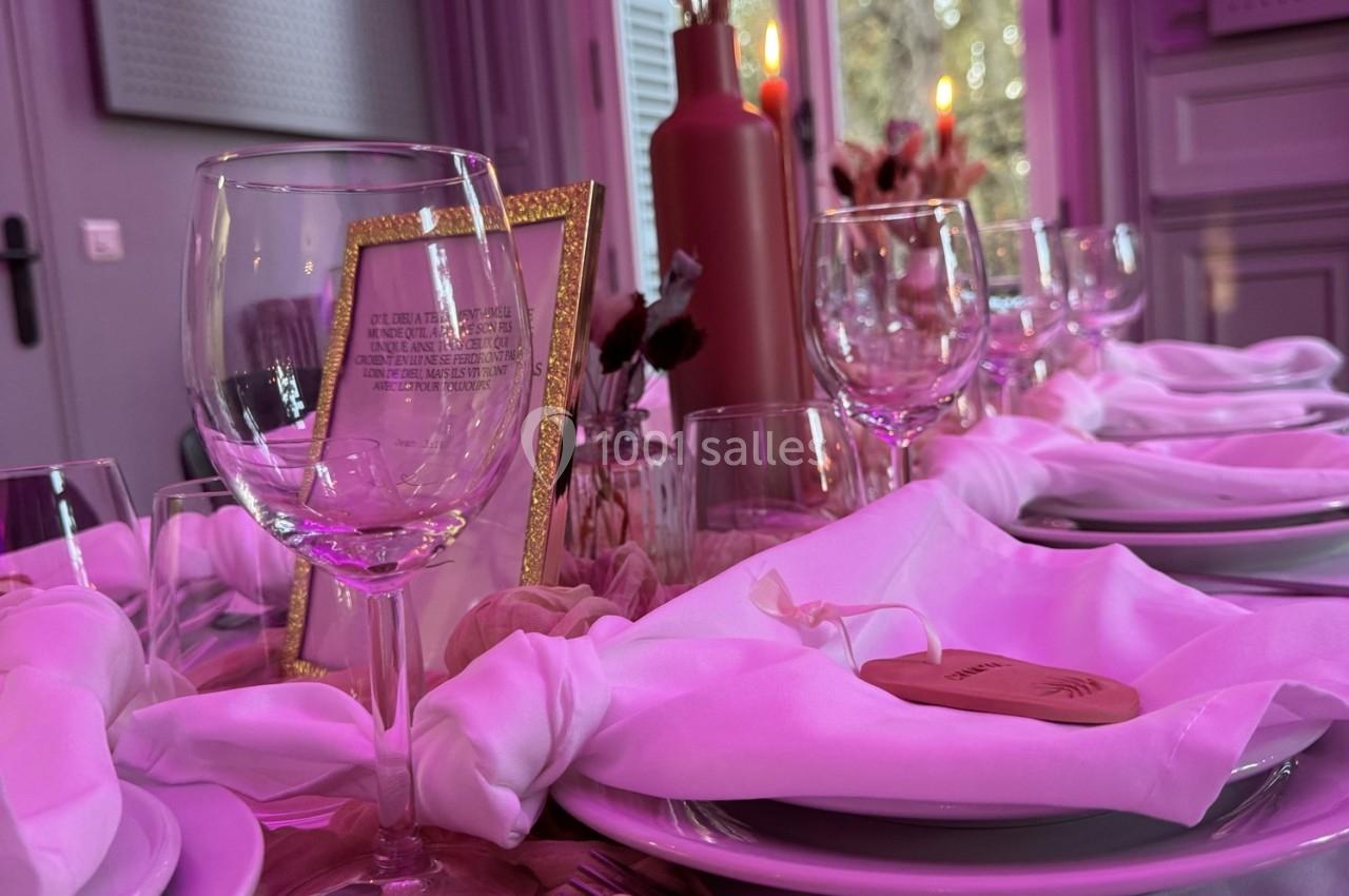 Table dressée avec des verres, serviettes blanches nouées et décorations roses dans une ambiance lumineuse tamisée.