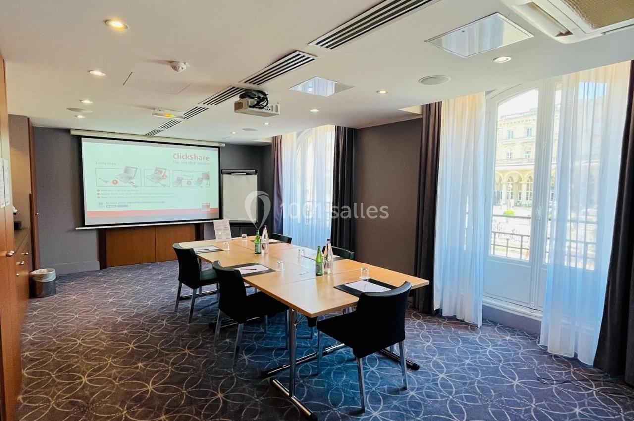 Salle de réunion lumineuse avec table rectangulaire, chaises noires, écran de projection et grandes fenêtres.