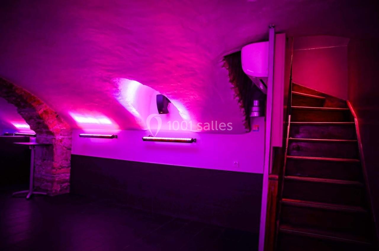 Salle voûtée éclairée par des lumières roses avec un escalier en bois sur la droite.