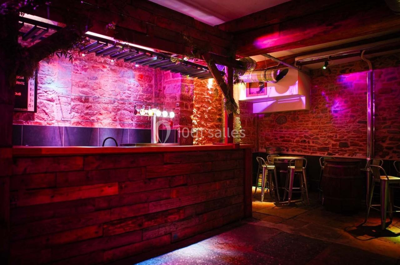 Bar en bois avec éclairage coloré, murs en pierre apparente et quelques tables hautes en métal.